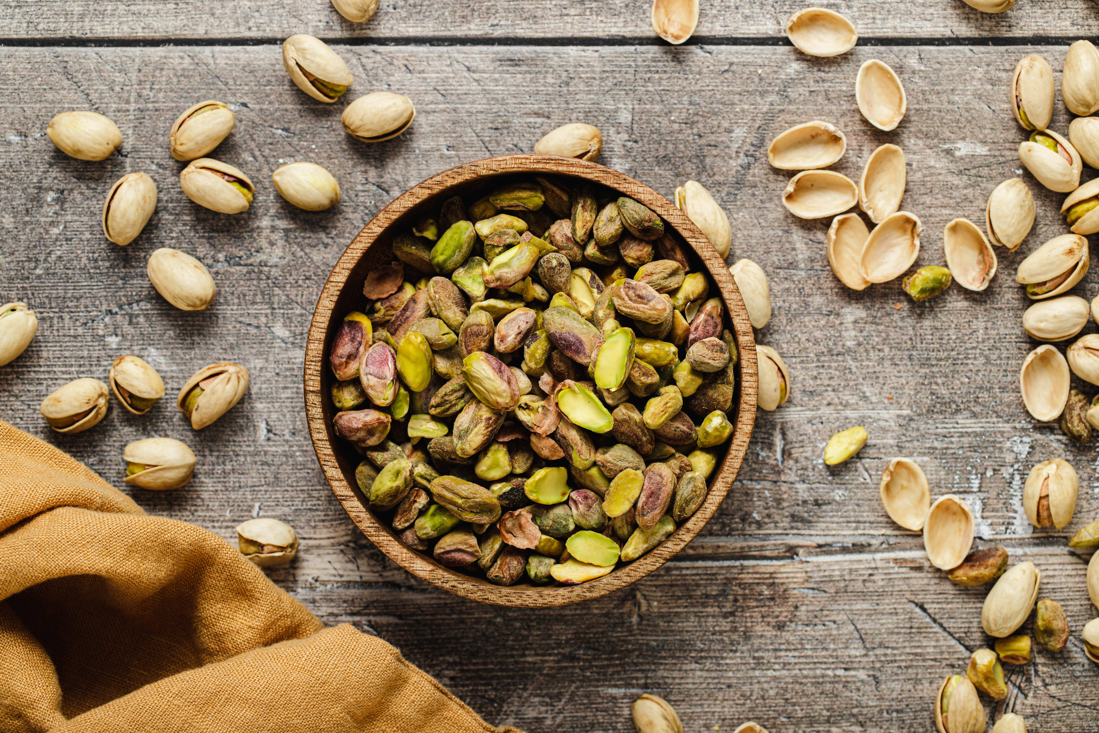 top-down pistachios in a wooden bowl with shells on wooden surface